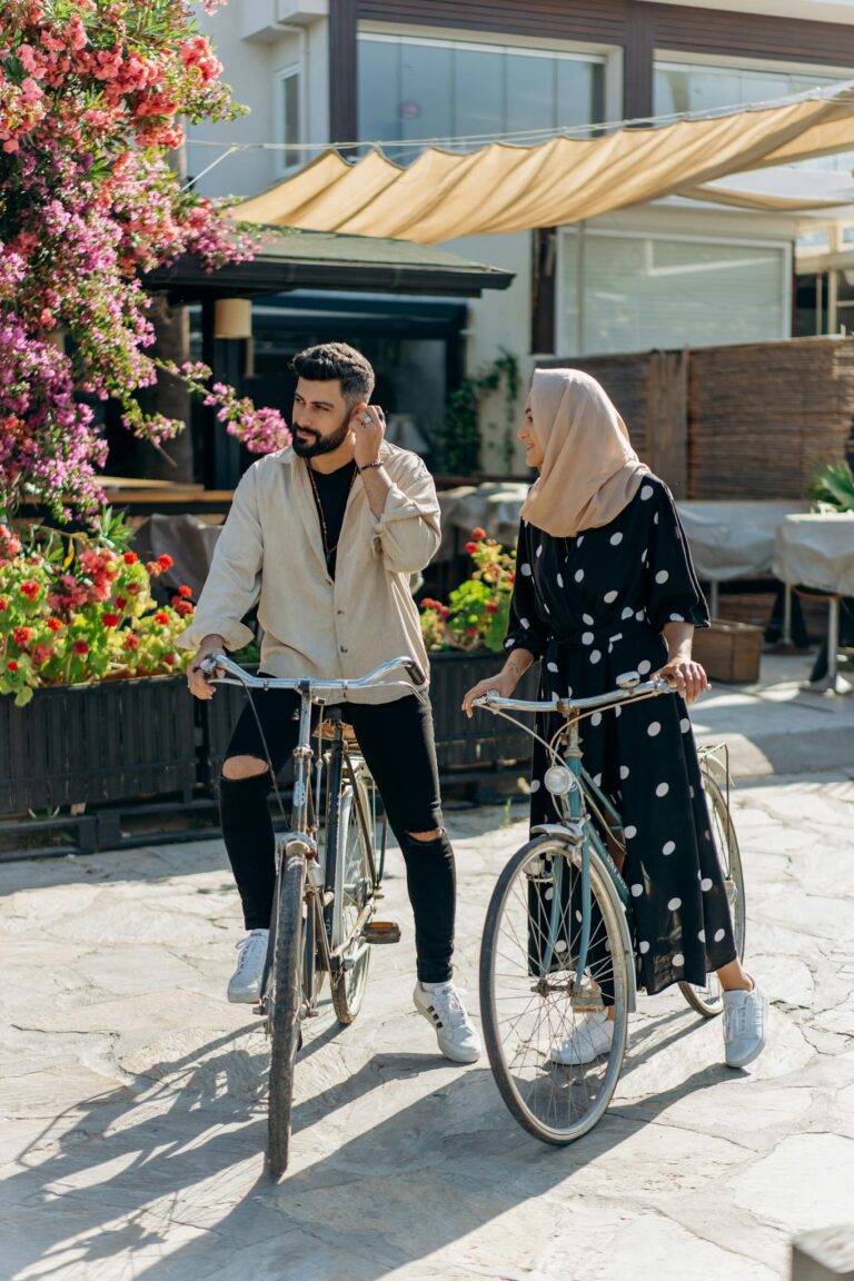 A man and woman in stylish attire biking together under sunny skies with floral surroundings.